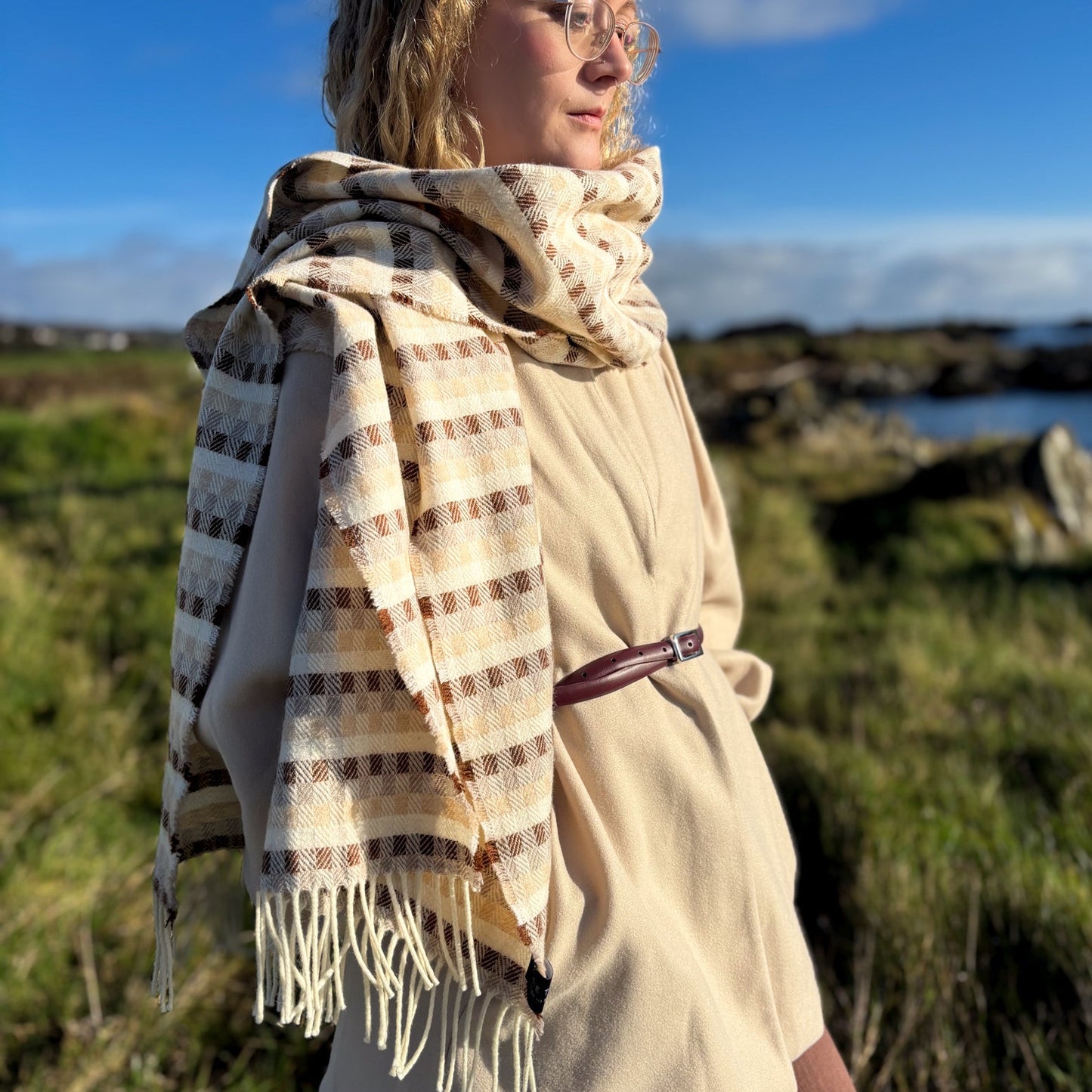 Woman wearing a beige coat and plaid scarf standing in a grassy outdoor setting with a body of water and rocks in the background.