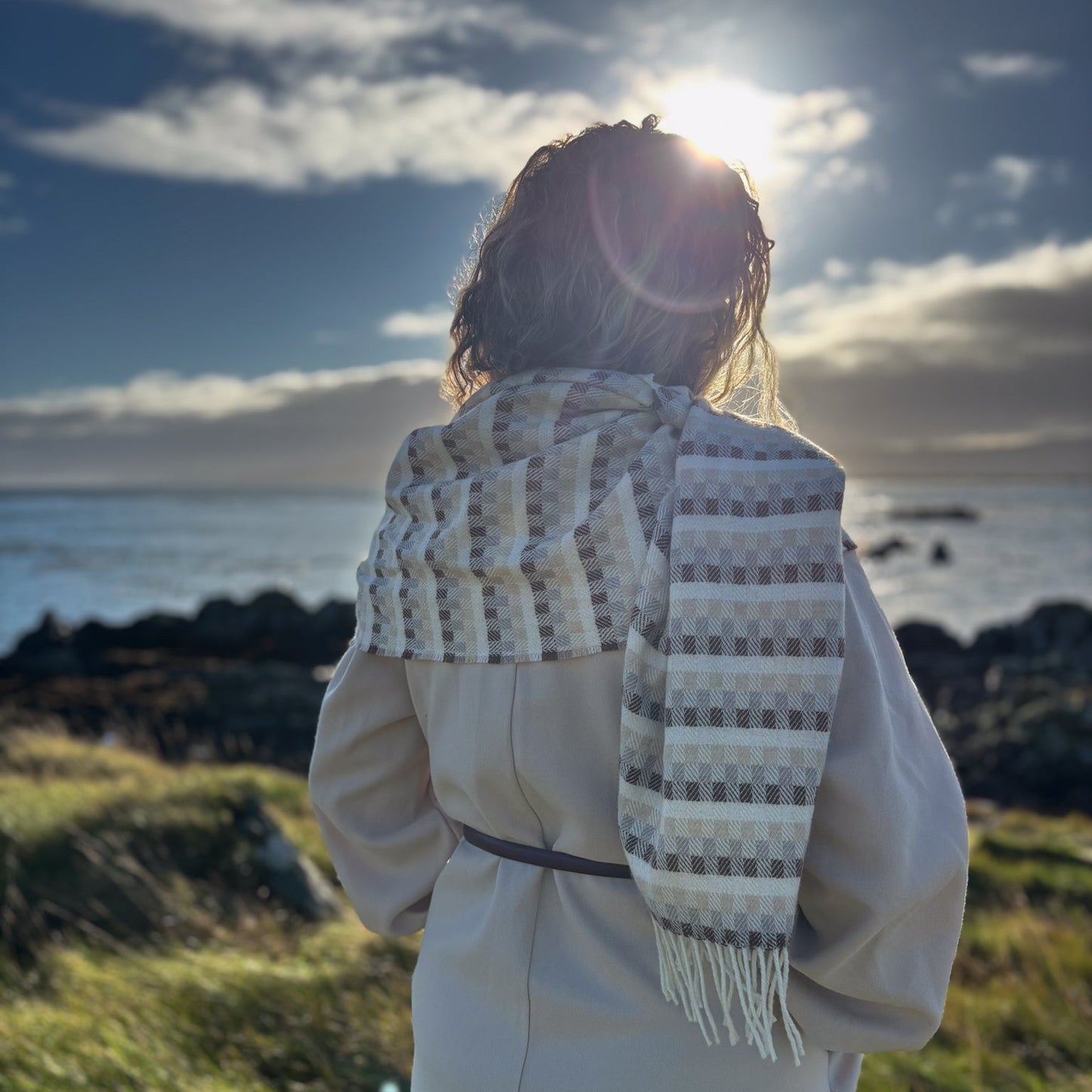 Person wearing a patterned scarf standing by the ocean with the sun in the background