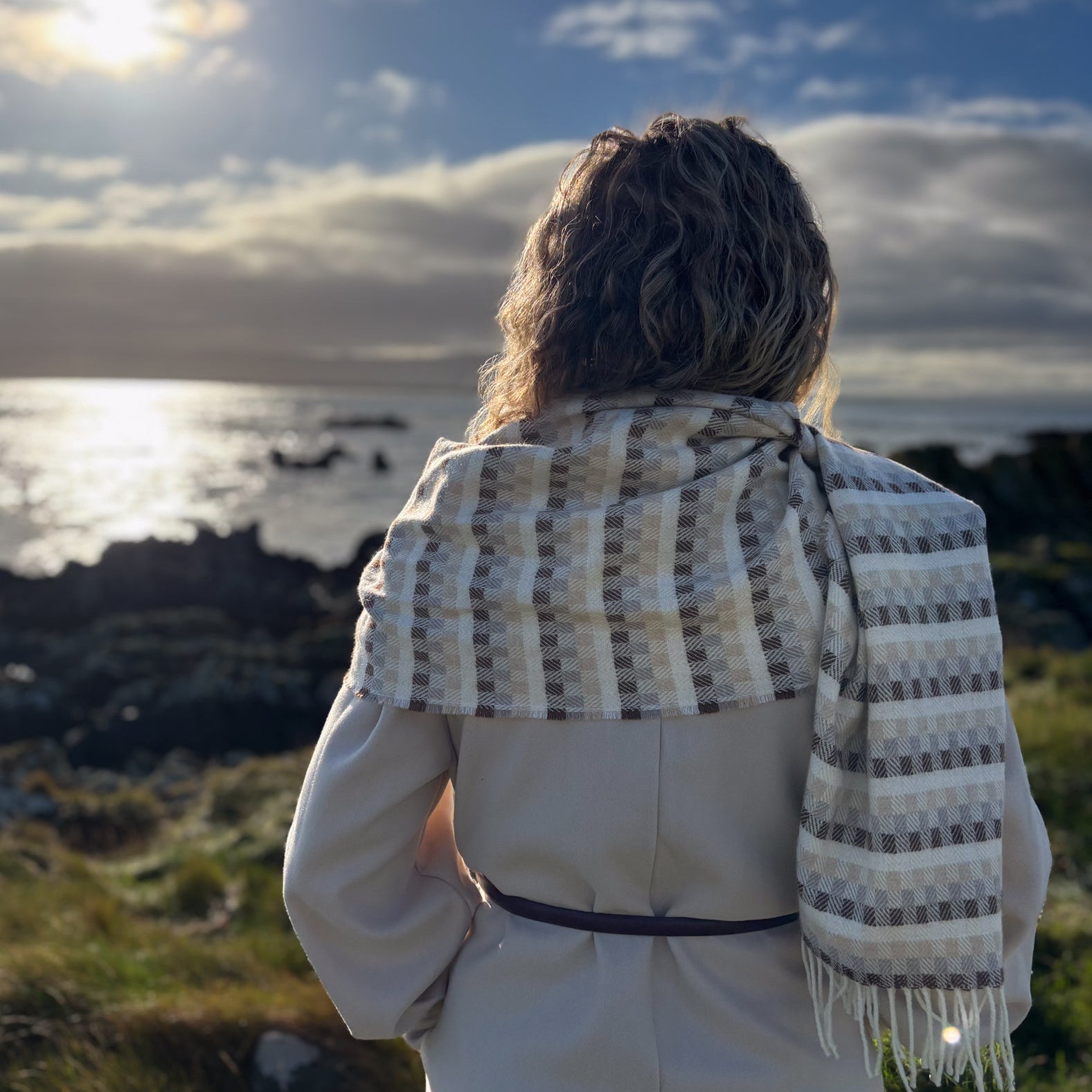 Person wearing a patterned tweed scarf standing on a coastal landscape with the sun shining.