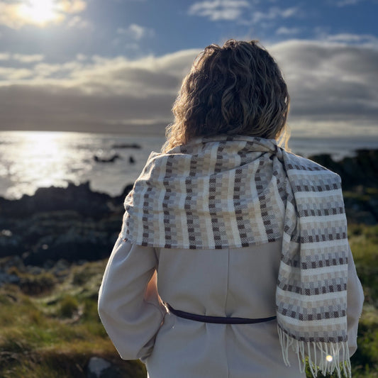 Person wearing a patterned tweed scarf standing on a coastal landscape with the sun shining.