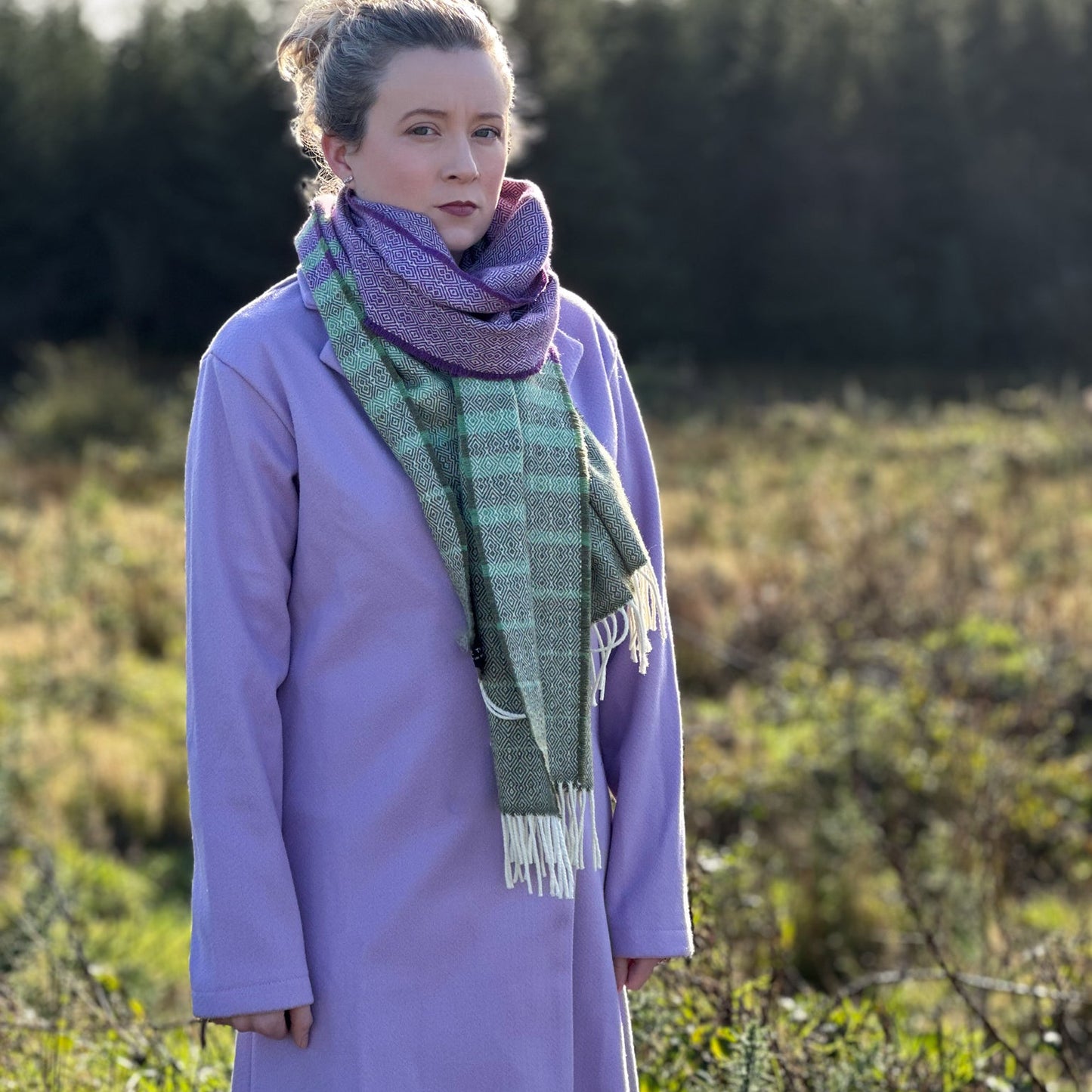 Person wearing a purple coat and green scarf standing in a field with trees in the background
