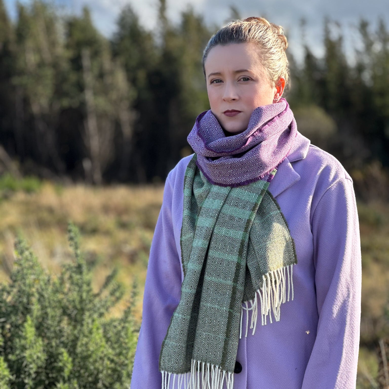 Person wearing a purple coat and green scarf standing in a natural setting with trees and sky.