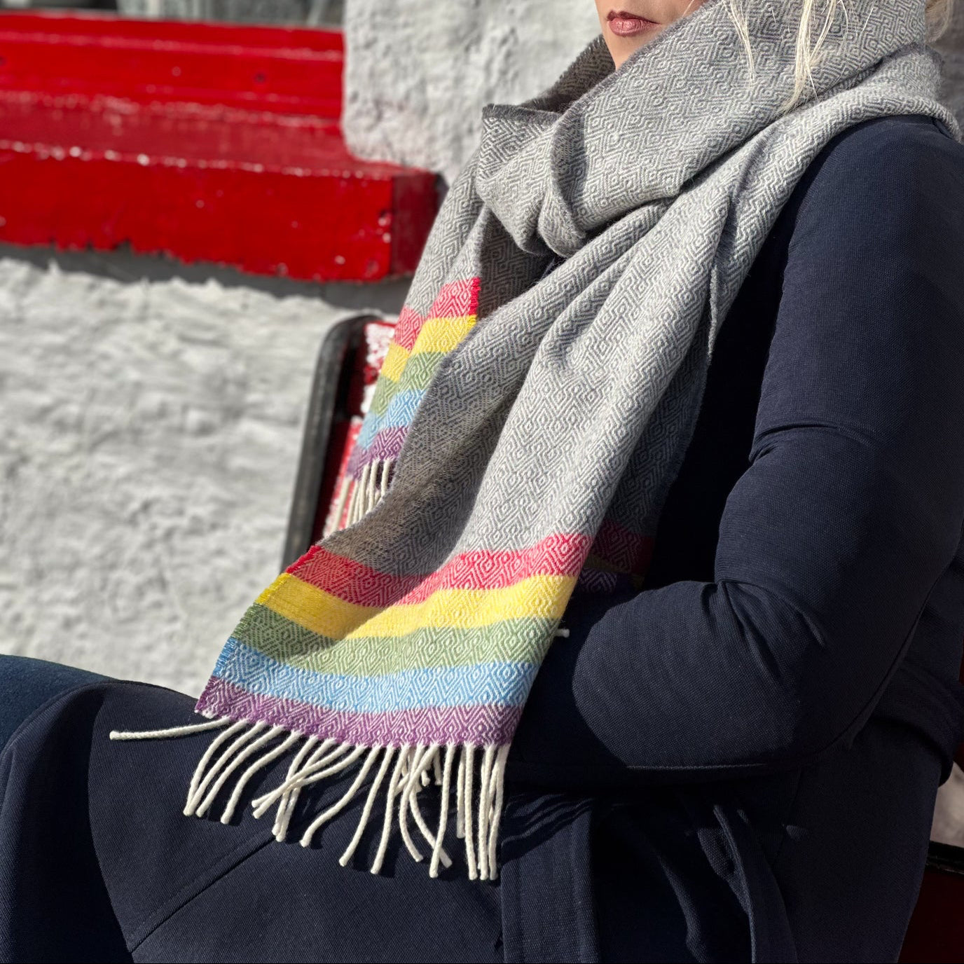 Woman wearing a gray scarf with a colorful pattern, sitting against a white wall.