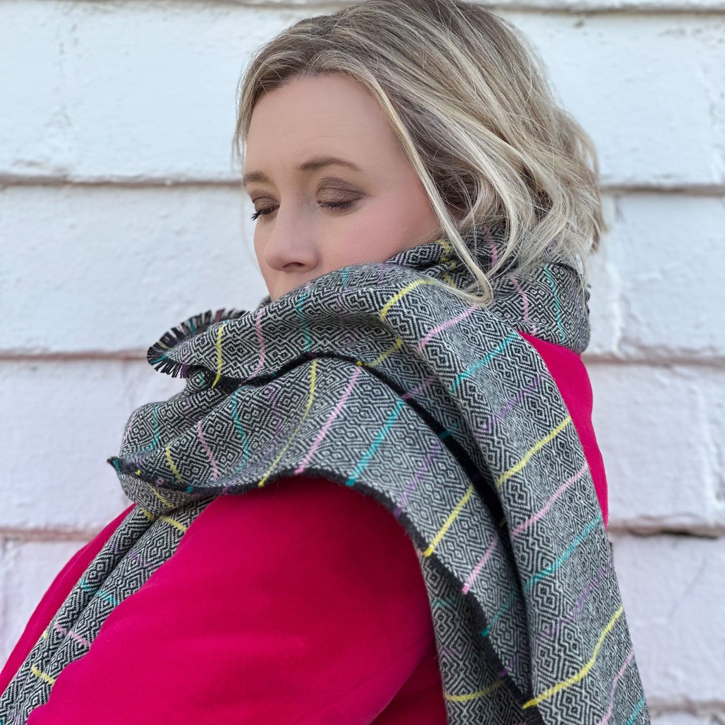 Woman wearing a plaid scarf with fringe over a pink jacket against a white brick wall.