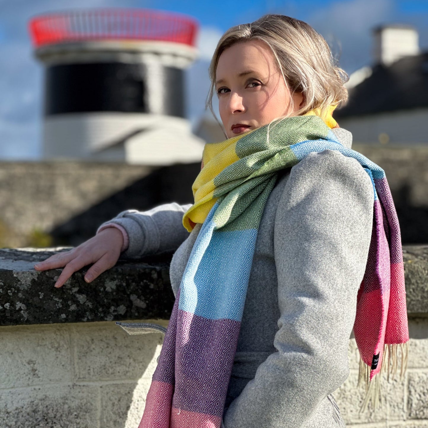 Woman wearing a colorful scarf and gray coat standing on a stone wall with a scenic background.
