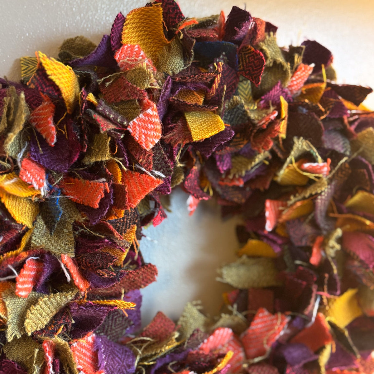 A handmade autumn-themed fabric wreath made from layered, frayed burlap strips in warm fall colors including orange, yellow, purple, red, and brown, shown against a plain off-white background.