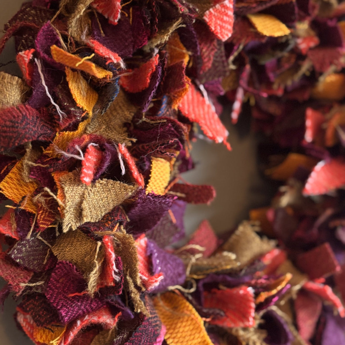 A close-up of a handmade autumn fabric wreath made from layered burlap and textured ribbon strips in fall colors including orange, red, purple, brown, and yellow.