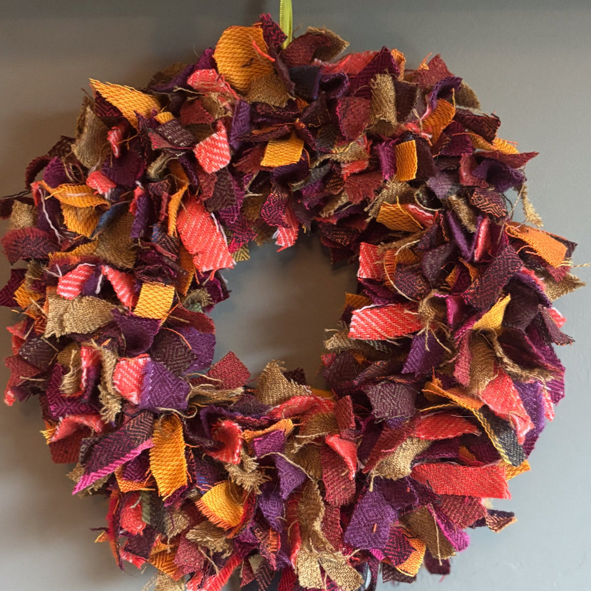 A handmade autumn-themed fabric wreath made from layered ruffled strips of orange, red, purple, brown, and tan burlap, hanging on a dark wooden door against a gray wall.