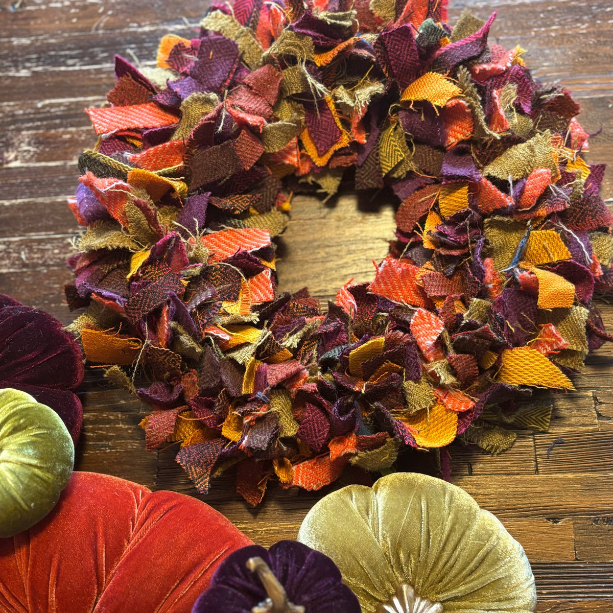A rustic autumn-themed fabric wreath made from layered shibori fabric strips in fall colors, displayed on a dark wood surface alongside velvet pumpkins.