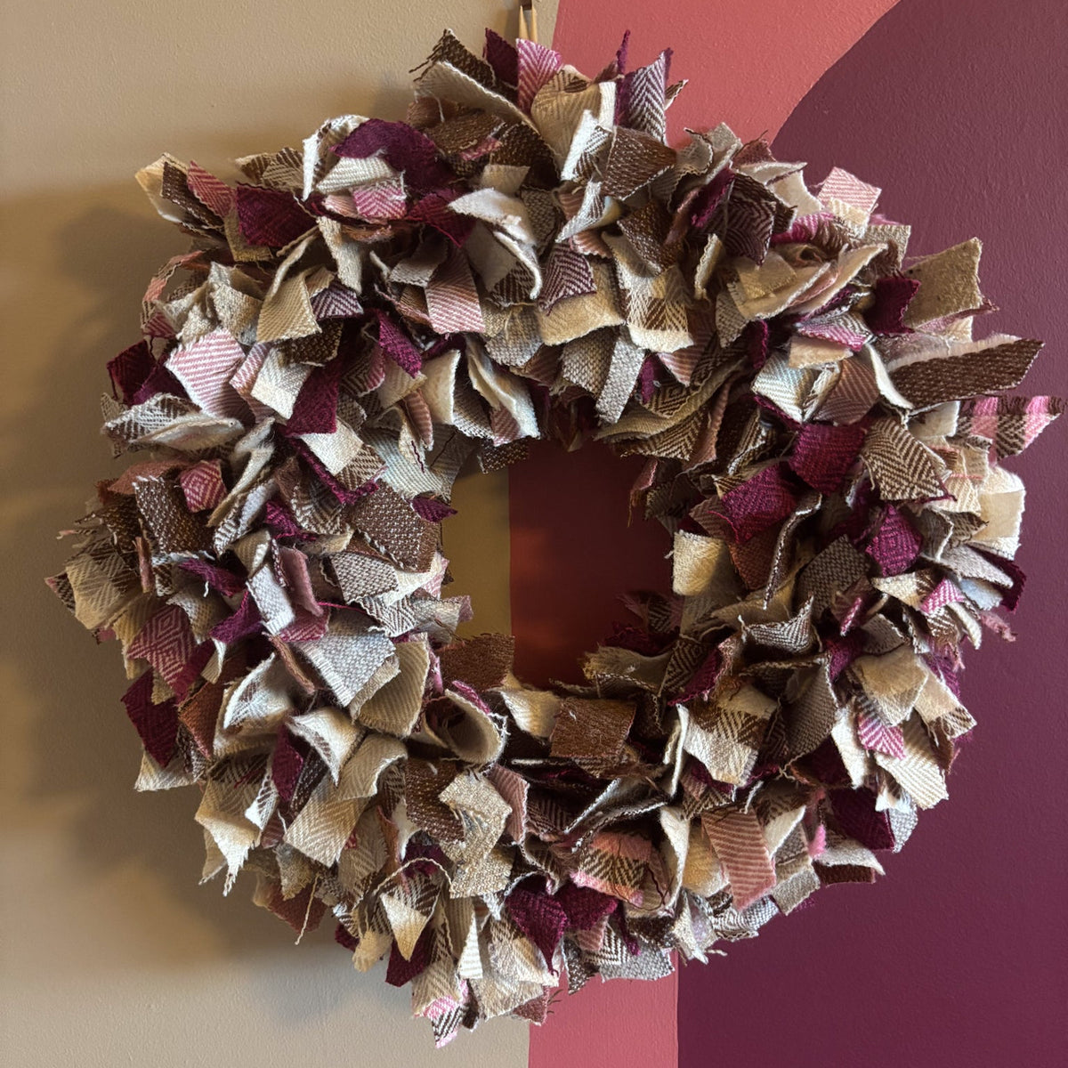 A handmade fabric wreath made from layered burlap and ribbon strips in beige, brown, and deep burgundy, hanging on a wall with a painted background of beige, pink, and dark purple sections.