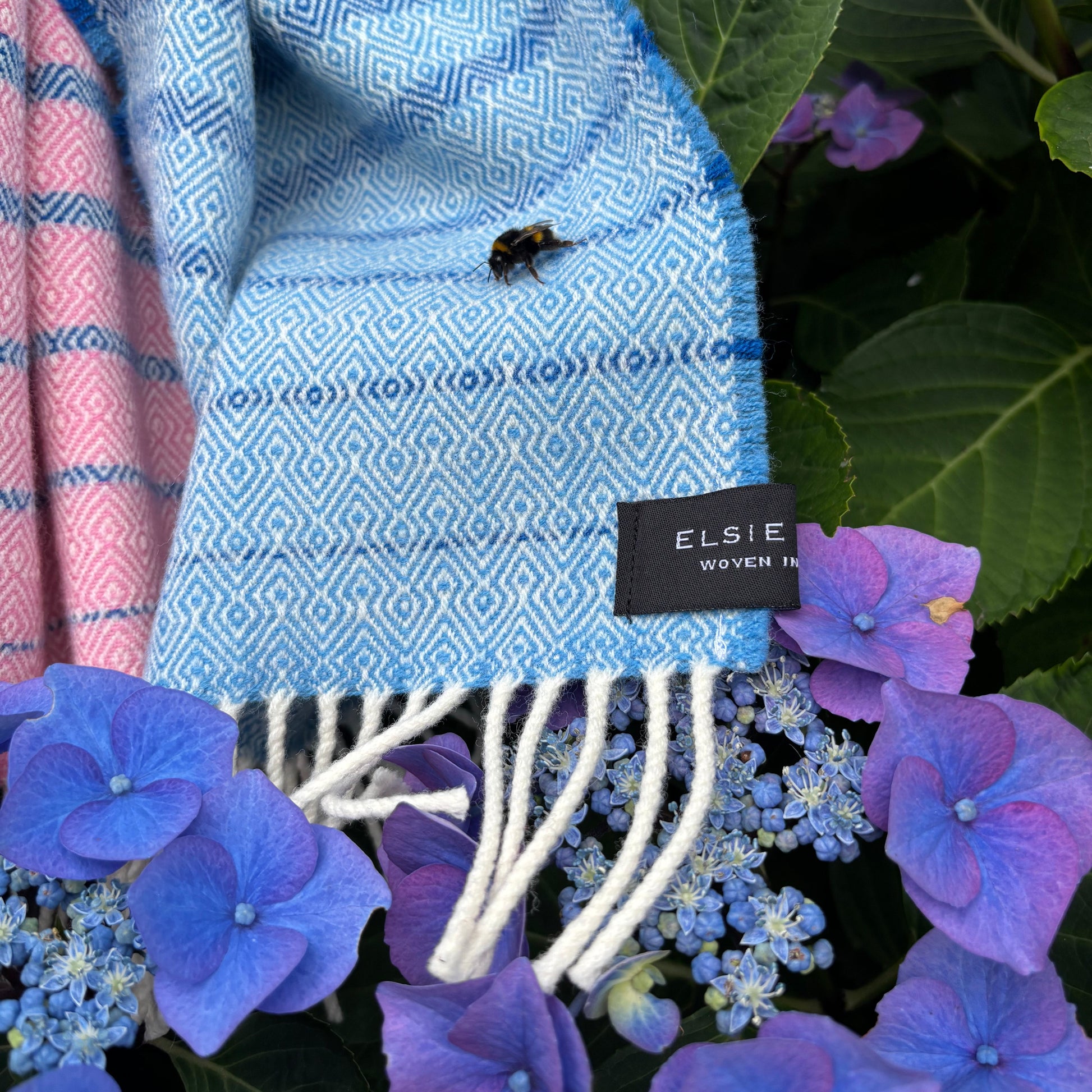 A folded light blue woven textile with a geometric pattern and white fringe, laid among green leaves and purple flowers. A black fabric tag reads ELSIE.