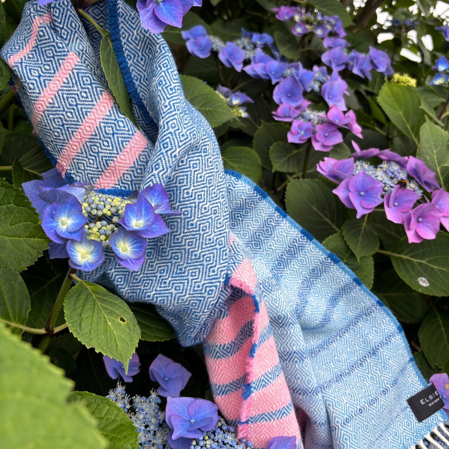 A light blue and pink woven scarf draped over purple hydrangea flowers and green leaves. The scarf features a geometric pattern on one side and pink stripes on the other, with white fringe at the top edge.