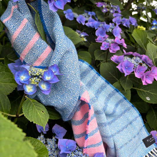 A light blue and pink woven scarf draped over purple hydrangea flowers and green leaves. The scarf features a geometric pattern on one side and pink stripes on the other, with white fringe at the top edge.