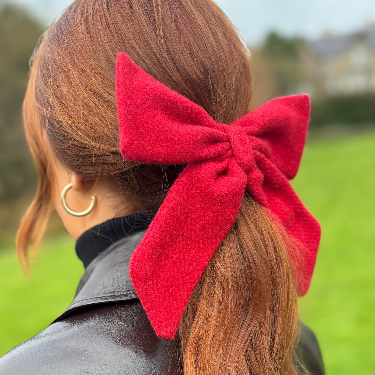 Person with red hair tied in a ponytail with a large red tweed bow, wearing a black leather jacket, standing in a grassy field.