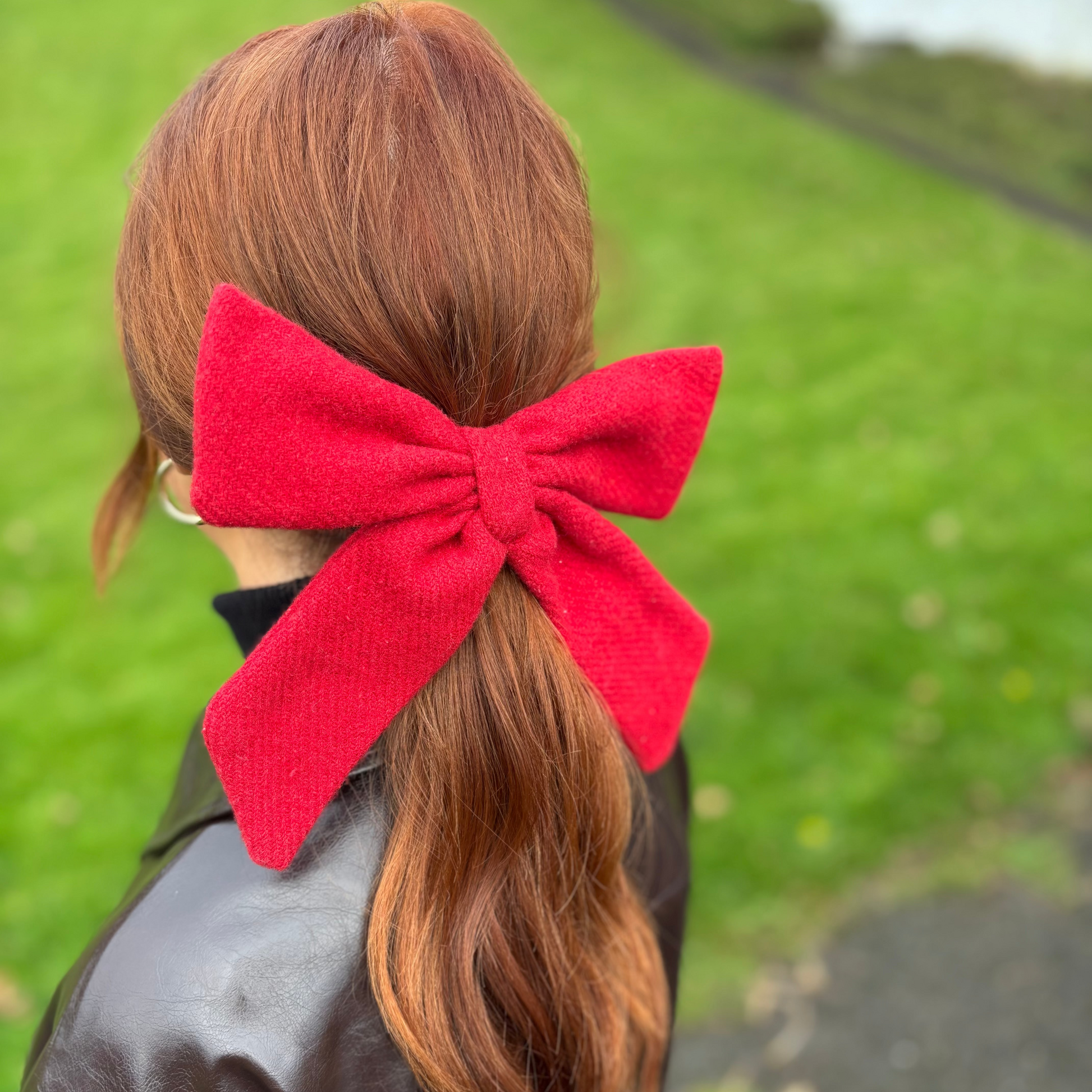 Person with red hair tied in a ponytail with a large red bow, standing outdoors.